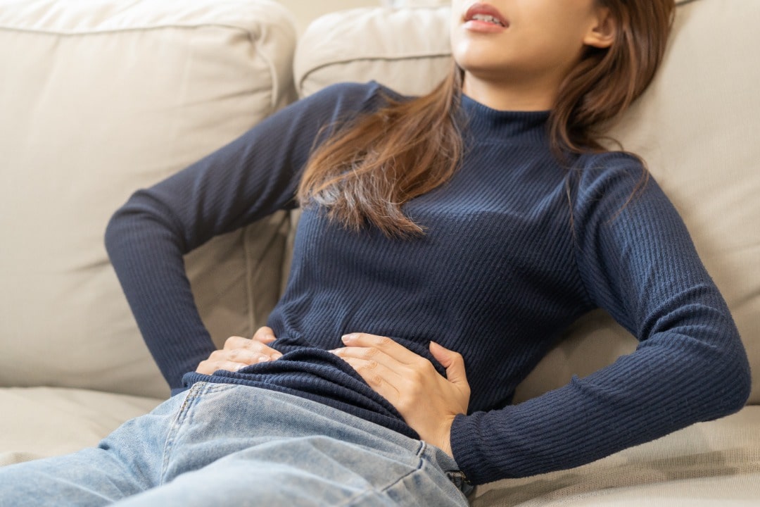 Woman sitting back on a couch hold her stomach in pain.