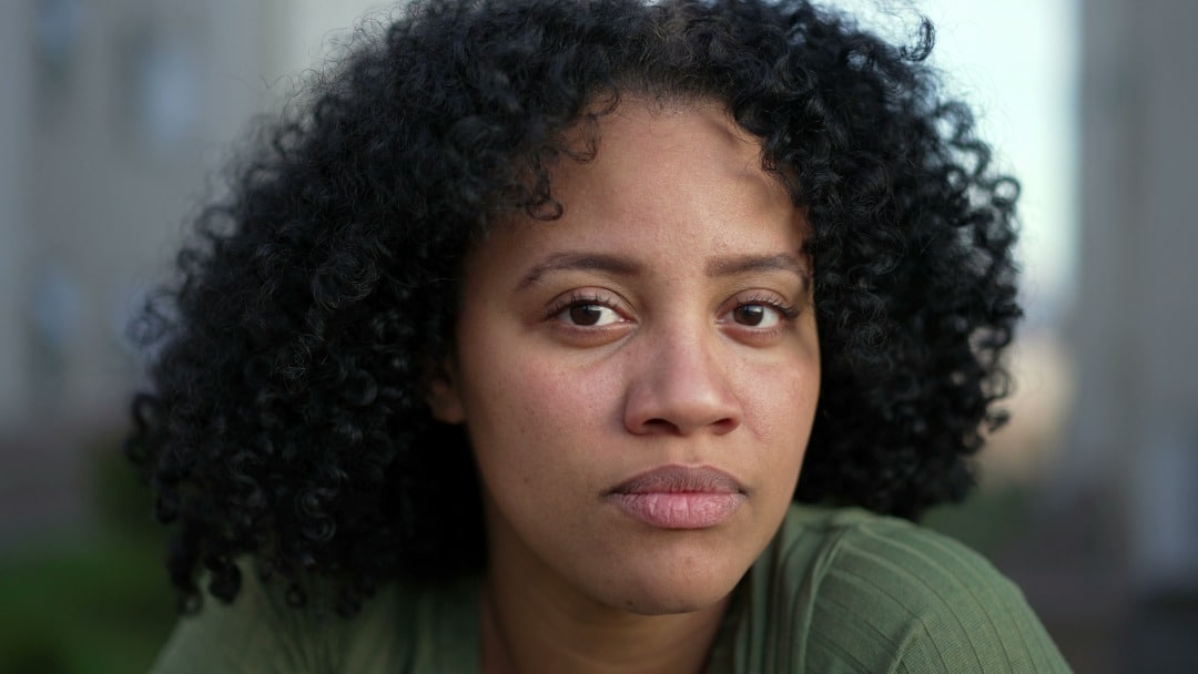 A Black woman in a green shirt looks at the camera with a serious look on her face.